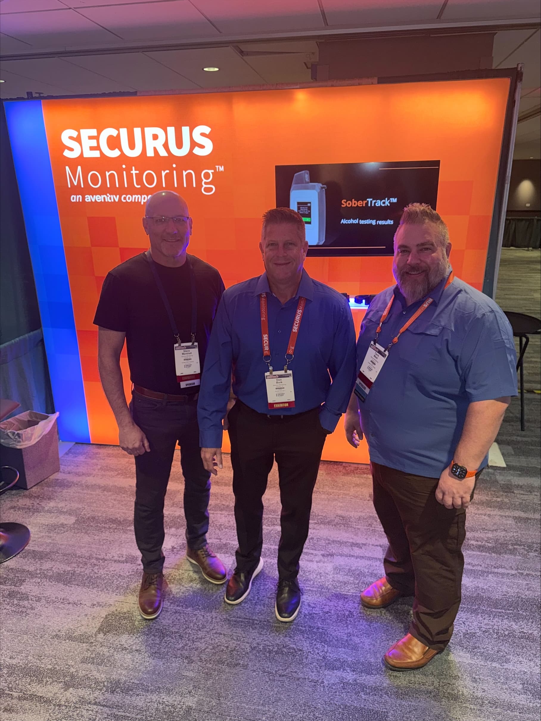 The image shows three men standing in front of a large orange and blue display for Securus Monitoring, a company that provides monitoring services.