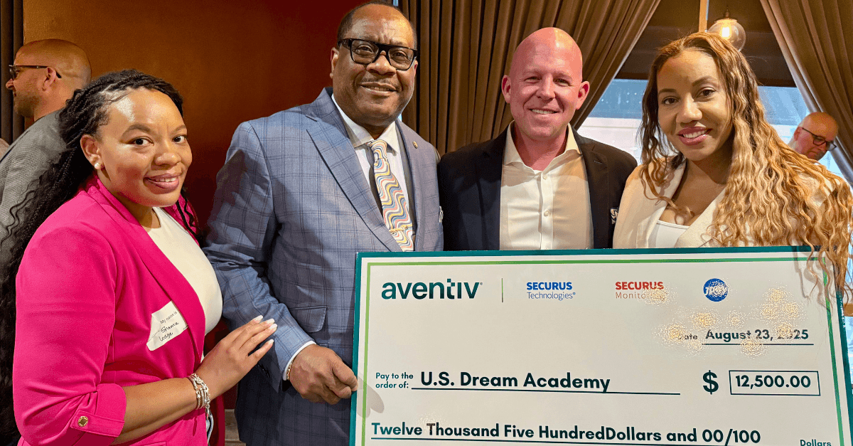 The image shows four individuals, two men and two women, posing together and holding a large check from Aventiv, a security company, presented to the U.S. Dream Academy.